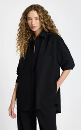 Camisa negra de tejido de punto con corte recto y ligeramente holgado. Presenta cuello estructurado, cierre frontal con botones, mangas largas con puños, bolsillo de parche delantero y bajo redondeado.