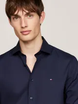 Camisa de manga larga para hombre, color azul marino, de corte slim fit, confeccionada en popelina con tecnología flex que incluye algodón orgánico. Presenta cuello con botones y el logo de Tommy Hilfiger bordado en el pecho.