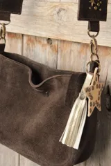 Bandolera confeccionada en cuero de gamuza marrón, con cierre superior. Presenta un llavero colgante con forma de estrella revestido en tachas metálicas y tiras flecadas en tono dorado.
