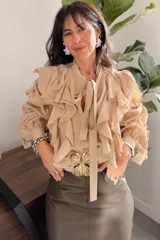 Camisa color camel con volados verticales en el frente y mangas largas abullonadas con puños ajustados. Presenta un lazo decorativo en el cuello y cierre frontal con botones.
