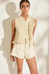 Top tejido sin mangas color beige con hilos de lurex, escote redondo y cierre frontal con tiras decorativas y botones dorados. Presenta detalle de volados en el ruedo.