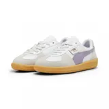 Championes Puma modelo Palermo LTH, con diseño de perfil bajo, cuerpo de cuero blanco, detalles en gamuza gris y franja lateral en color lila. Cuenta con suela de goma color caramelo y logo dorado en el lateral.