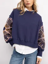 Buzo de cuello redondo en color azul marino, con diseño de bordado floral XL en tonos naranja y rosado en ambas mangas. Presenta un corte holgado con puños y ruedo acanalados.