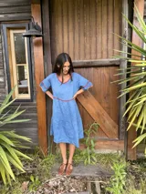 Vestido midi de lino color azul, con escote en V, mangas cortas y falda amplia de corte escalonado. Presenta detalles de costuras y bordes en tono rojizo contrastante.