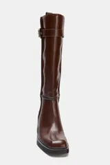 Bota alta de cuero sintético color marrón oscuro, con caña recta y punta cuadrada. Presenta pespuntes a contraste en las uniones y un detalle de correa con hebilla metálica en la parte superior. Posee un tacón de bloque de altura media.