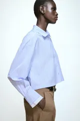 Camisa corta de corte recto con estampado de rayas verticales en tonos azul y blanco. Presenta cuello solapa, cierre frontal con botones, bolsillo de vivo en el pecho y puños anchos en color blanco a contraste.