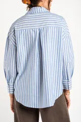 Camisa de algodón de corte holgado y diseño a rayas verticales en color celeste y blanco, con cuello camisero, cierre frontal de botones, bolsillo aplicado en el pecho y mangas largas con puños abotonados.