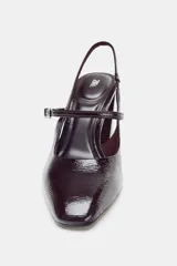 Zapato tipo mercedita destalonado de charol color borgoña oscuro, con punta cuadrada. Posee una tira delantera con hebilla metálica y una tira trasera elástica. El tacón es alto y de bloque.