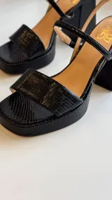 Sandalia de tacón alto color negro con textura de piel de serpiente, plataforma delantera, punta cuadrada y tira fina en el tobillo con hebilla.