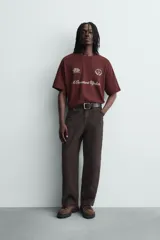 Camiseta de manga corta y cuello redondo, color granate oscuro, con estampado frontal en blanco que incluye texto y un escudo circular.