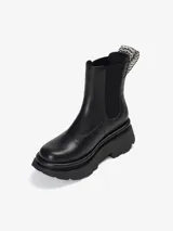 Bota tipo Chelsea de caña media confeccionada en cuero de oveja color negro. Presenta paneles elásticos laterales, tirador trasero con detalle tejido y suela chunky de goma con relieve.