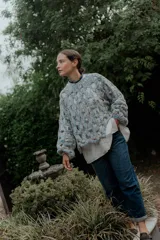 Buzo de tejido de punto calado con diseño de red, cuello redondo y mangas largas abullonadas. Presenta una textura jaspeada en tonos gris y blanco.