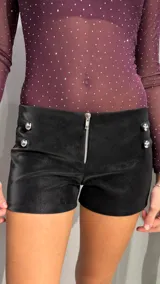 Short de tiro bajo confeccionado en material engomado efecto cuero color negro. Presenta cierre frontal metálico y detalle de botones plateados decorativos en ambos laterales de la cintura.