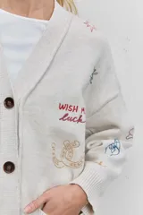 Cardigan de punto azul con cuello en V, cierre frontal de botones y bolsillos delanteros. Presenta bordados decorativos con motivos variados y frases en colores contrastantes.