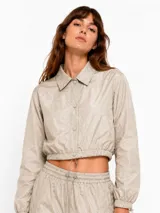 Campera corta de nylon color beige, con cuello camisero, cierre frontal con botones a presión, mangas largas con puños elásticos y cordón ajustable en la cintura.