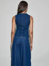 Chaleco de jean azul oscuro, sin mangas, con cuello redondo y cierre frontal de botones metálicos dispuestos en línea curva hacia el hombro izquierdo.