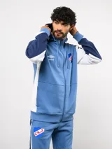 Campera deportiva Umbro del Club Nacional de Football, con cierre frontal, capucha ajustable con cordones y diseño en bloques de color azul, celeste y blanco. Presenta el logo de la marca y el escudo del club bordados en el pecho.