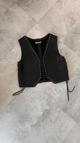 Chaleco de cuero de diseño clásico, con escote en V y detalles de tachas metálicas en los bordes delanteros. Cuenta con cordones laterales ajustables en contraste.