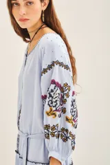 Vestido corto de bambula color celeste, con escote redondo, mangas tres cuartos abullonadas y falda con volado en el ruedo. Presenta bordados decorativos en el frente y las mangas, y cuenta con un cinturón de la misma tela para ajustar a la cintura.