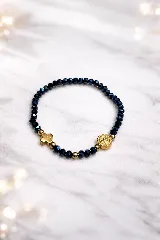Pulsera elástica compuesta por cuentas facetadas de color azul oscuro brillante, intercaladas con cuentas redondas doradas. Incluye un dije de cruz dorada y un dije redondo con una medalla religiosa dorada.