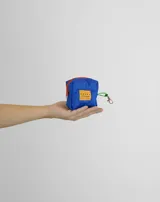 Monedero pequeño de forma cúbica en color azul, con cierre superior rojo y gancho metálico lateral con cinta verde. Incluye una etiqueta rectangular naranja con el logo de la marca en el frente.