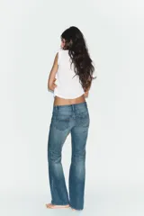 Jeans de tiro bajo con corte bootcut, confeccionados en denim con efecto lavado. Presentan costuras verticales marcadas en el frente, cierre frontal con cremallera, gancho metálico y detalle de trabilla con botones en la cintura.