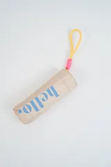 Paraguas plegable color beige con la palabra 'hello.' estampada en azul. Incluye una manija de transporte de silicona amarilla con detalle de hilo rosado.
