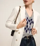 Blazer blanco de corte sastre con botones dorados y cuello solapa. Se combina con un sweater de punto sin mangas (chaleco) con patrón argyle en tonos gris y azul marino, y un pantalón de jean oscuro.