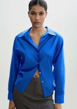 Camisa de corte recto y tejido satinado color azul eléctrico, con cuello camisero, manga larga con puños abotonados y cierre frontal de botones.