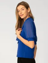 Blusa de color blanco con escote en V y detalle de lazo frontal. Presenta mangas cortas de gasa con terminación en puño.