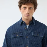 Camisa de denim azul con diseño de rayas verticales finas y blancas, manga larga con puños ajustables, cierre frontal de botones y dos bolsillos de parche con solapa en el pecho.