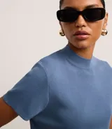 Blusa de punto azul medio, ajustada al cuerpo, con cuello alto y mangas cortas. El tejido presenta una textura de malla fina.