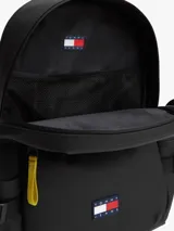 Mochila negra de poliuretano con compartimento principal y cierre de doble cremallera con tiradores amarillos a contraste. Cuenta con bolsillo frontal con cremallera, asa superior, correas ajustables y acolchadas, y parche engomado de Tommy Jeans en la parte delantera.