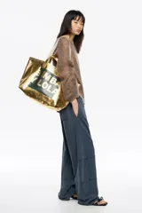 Bolso tipo tote de Bimba y Lola, color dorado con acabado metalizado y efecto arrugado. Presenta un diseño cuadrado con estampado central de la marca en un recuadro oscuro y doble asa de mano en color crema.