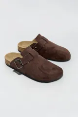 Zueco tipo slip-on de gamuza marrón, con tira y hebilla metálica regulable en el empeine, suela de corcho y base de goma negra.