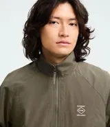 Campera rompeviento color verde militar, con cierre frontal de cremallera, cuello alto, puños y cintura elásticos. Presenta recortes delanteros y un pequeño logo estampado en el pecho.