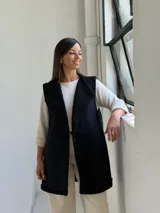 Chaleco largo de gamuza sintética color negro, con interior y bordes de corderito sintético al tono. Presenta un diseño abierto sin mangas, ideal para superponer sobre otras prendas.