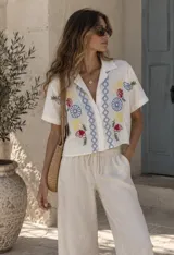 Camisa de manga corta color beige, con cuello solapa y cierre frontal de botones. Presenta bordados coloridos de flores y motivos geométricos en el frente.
