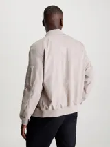 Campera tipo bomber de Calvin Klein en color beige, confeccionada en una mezcla de algodón y poliamida. Presenta cuello alzado, puños y bajo de punto canalé, cierre frontal de cremallera, bolsillos laterales de hendidura y un bolsillo adicional con cremallera en la manga izquierda con insignia de goma de la marca.