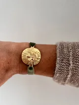 Pulsera con banda de terciopelo verde oliva y dije circular dorado con figura de paloma y rayos.