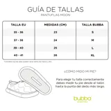 Pantuflas acolchadas de diseño tipo slip-on en color lila, con costuras onduladas y suela de goma blanca. Presentan el logo de la marca bordado en amarillo en la parte superior.