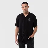 Camisa de manga corta en color negro con cuello tipo cubano y cierre frontal de botones. Presenta un pequeño estampado circular en el pecho con texto blanco.