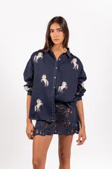 Camisa de corte clásico y calce amplio, color marfil, con cuello camisero, mangas largas y cierre frontal con botones. Presenta bordados coloridos de caballos en contraste distribuidos en la prenda.