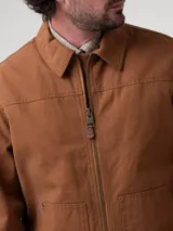 Chaqueta de canvas color marrón, con cierre frontal completo, cuello clásico y forro interior de sherpa. Presenta dos bolsillos frontales tipo parche y corte regular.