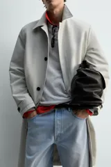 Trench coat de corte relajado, color gris claro, confeccionado en tejido técnico con mezcla de algodón. Presenta cuello solapa, manga larga con puño ajustable, bolsillos de doble vivo en cadera, bolsillo interior y abertura en el centro de la espalda. Cierre frontal con botonadura oculta.