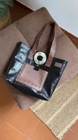 Tote bag de ecocuero negro con textura de reptil, que presenta un bolsillo frontal en tono marrón con remaches metálicos. Incluye manijas en color marrón y un detalle decorativo circular de guampa en la parte superior.