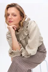 Blusa de lino color beige, con cuello alto fruncido y detalles de volados en el cuello y los puños elásticos. Presenta mangas largas abullonadas y diseño holgado.