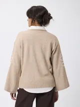 Sweater de tejido de punto color beige, con cuello redondo, mangas largas acampanadas y diseño bordado en color blanco en las mangas.
