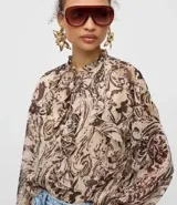 Blusa de chiffon con estampado paisley en tonos tierra, cuello alto con lazo frontal, volados decorativos y mangas largas con puños ajustados.
