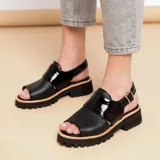 Sandalia de cuero vacuno color negro con diseño de punta abierta y talón descubierto. Presenta una tira ancha sobre el empeine en cuero charolado y una tira ajustable con hebilla metálica en el tobillo. Cuenta con una suela de goma tipo track con plataforma bicolor.